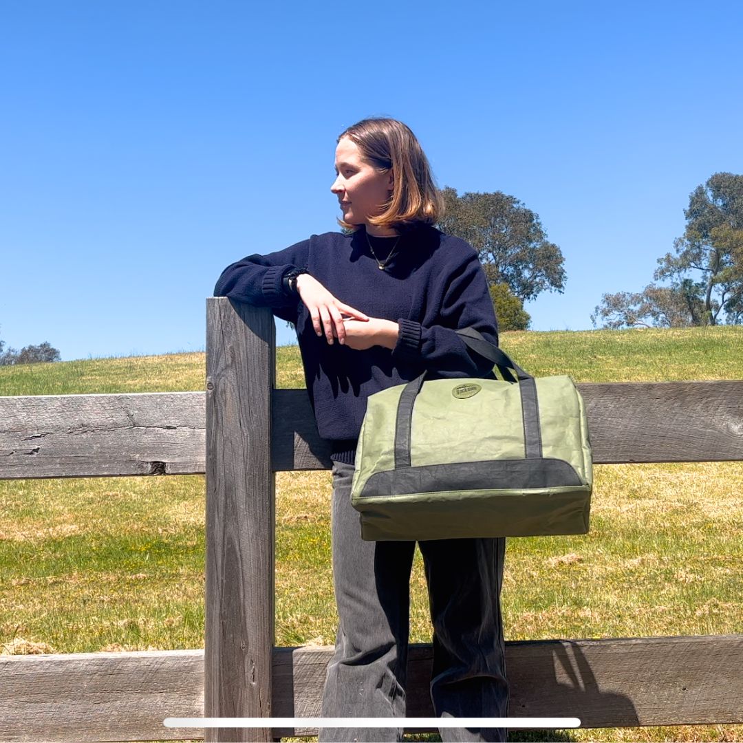 A girl stands holding the Joy Krasft Travel Bag while leaning on a fence looking to the side, 