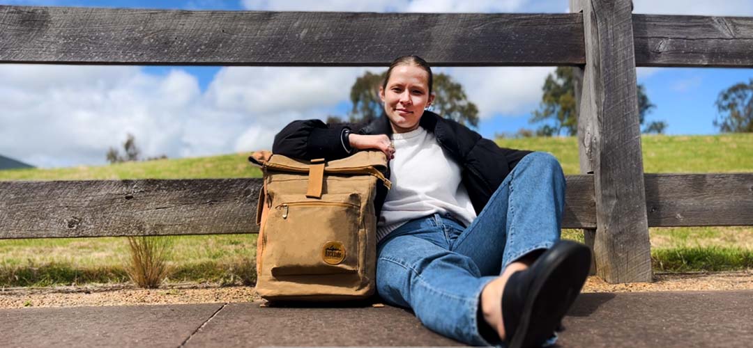 Courtney leaning on a wooden fence with a green background, holding the Rockside Northshore Kraft Paper Laptop Backpack under her right arm, showcasing the eco-friendly and stylish design of the backpack.