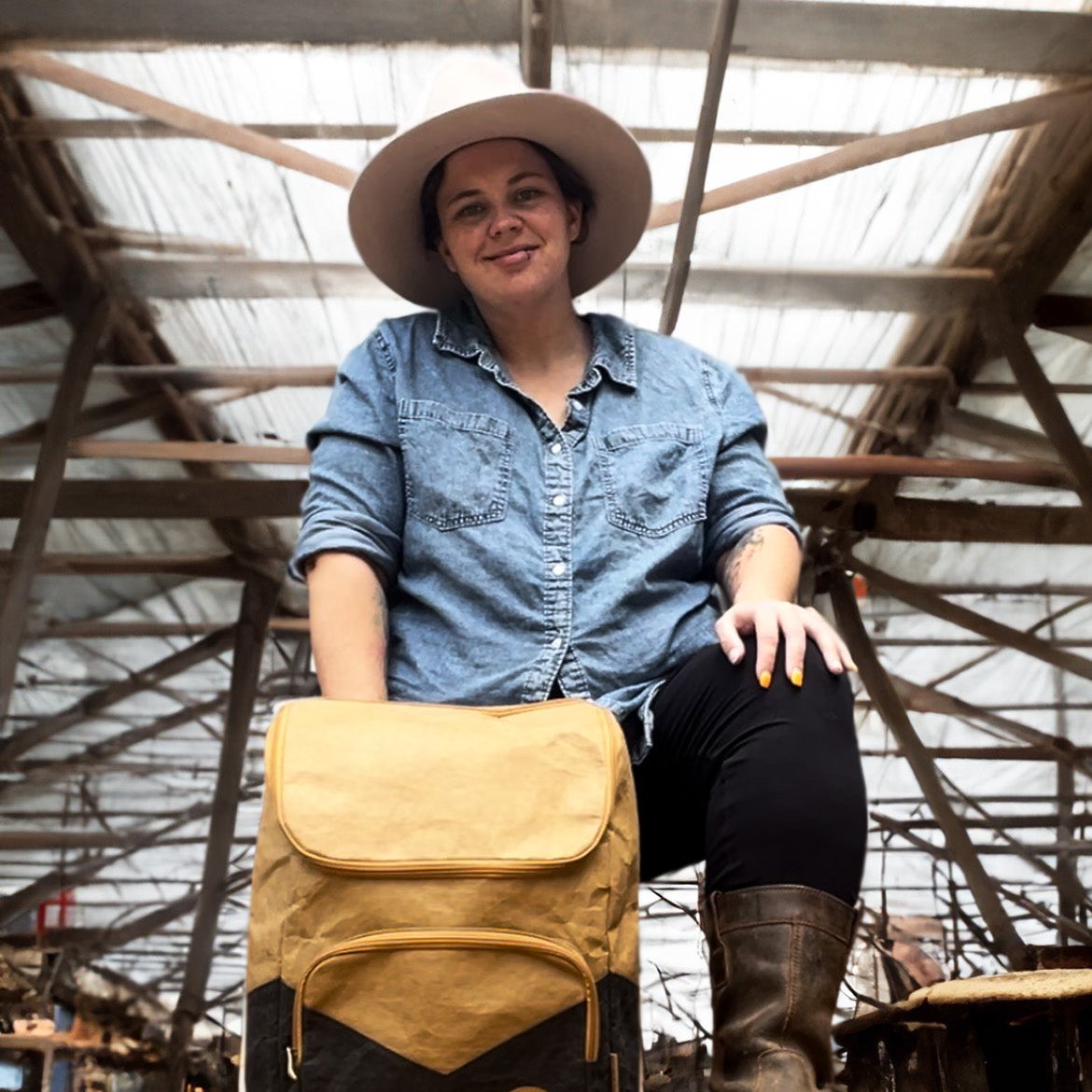 Rachael wearing a wide-brimmed hat stands above the Rockside Base Kraft Paper Laptop Backpack in front of a barn, showcasing the eco-friendly, durable design of the backpack