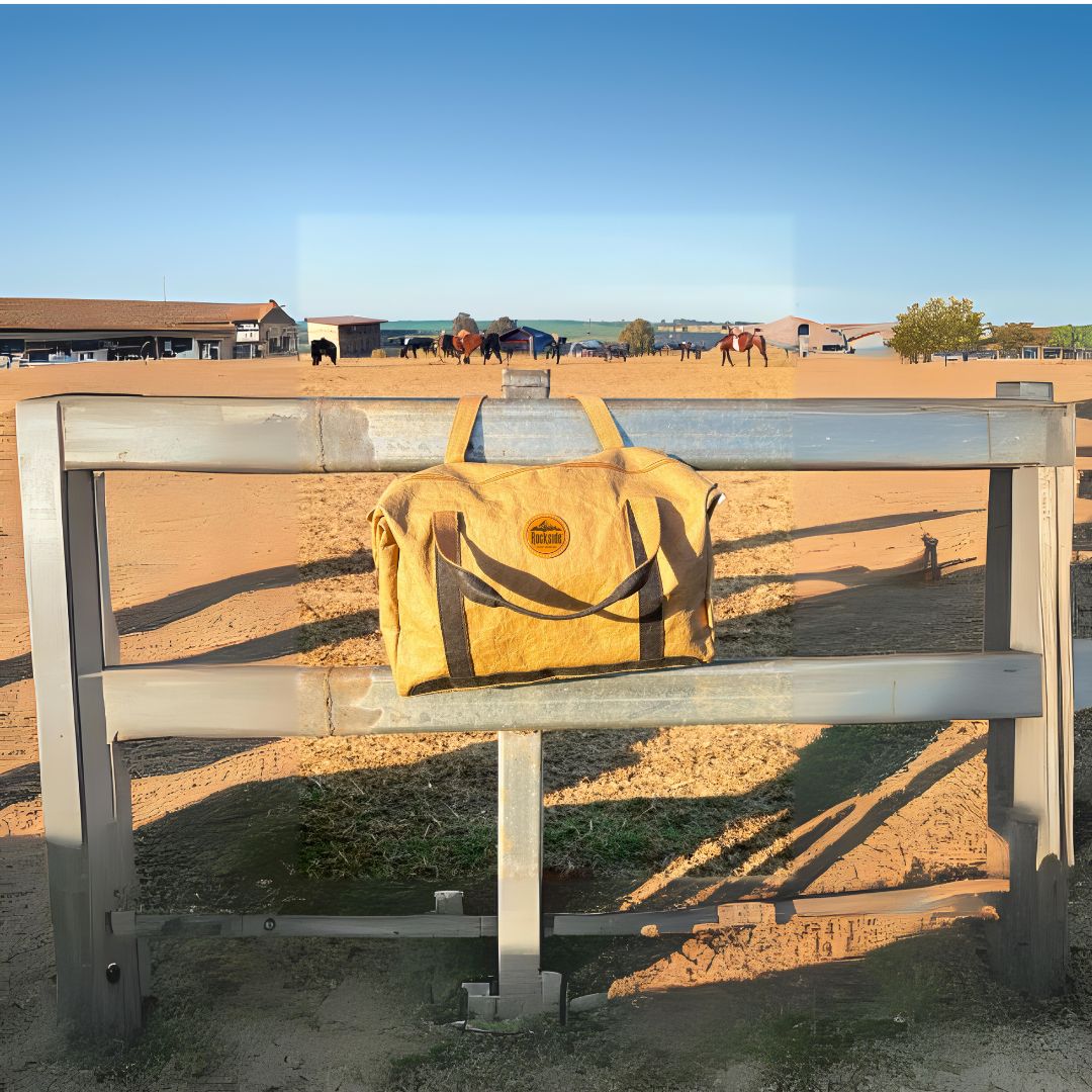 Rockside Joy Kraft Travel Bag resting on a fence in front of a horse paddock, with horses grazing in the background, showcasing the bag's durable and eco-friendly design